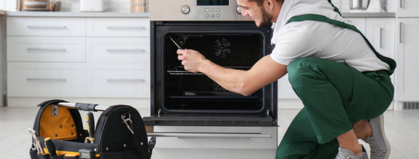 Repair man smiling as he reaches inside an open oven with his tool to repair an oven. An open toolbox sits by the open oven door in a white kitchen